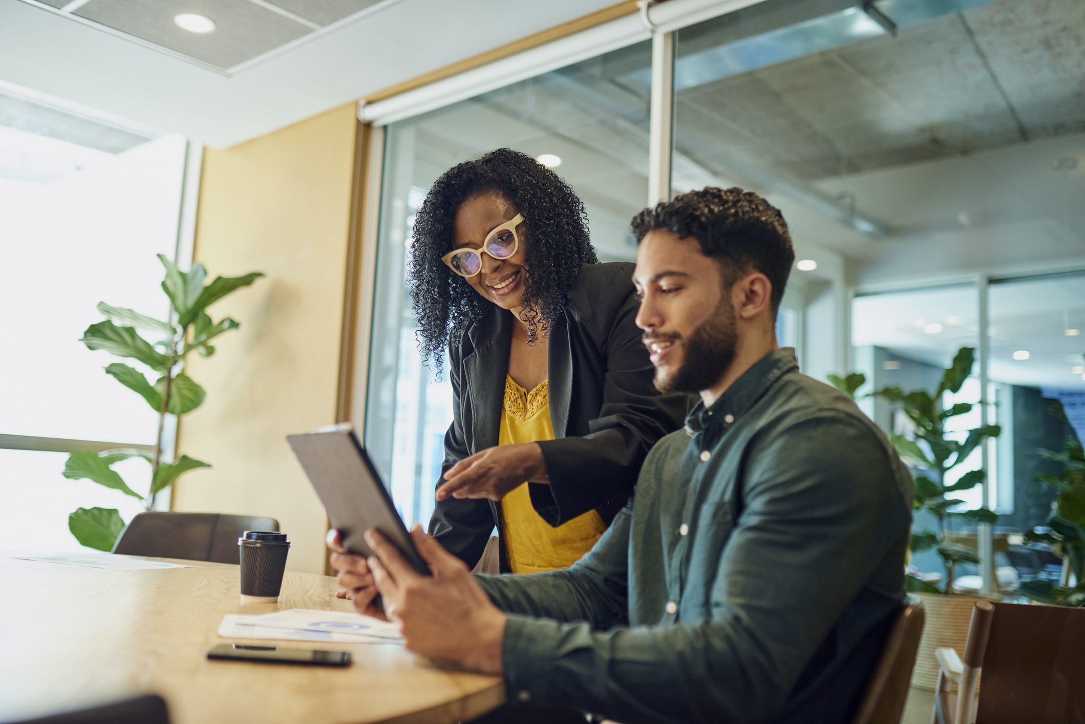 A smiling female coworker wearing glasses shows a tablet to her male colleague while sitting at a conference table.