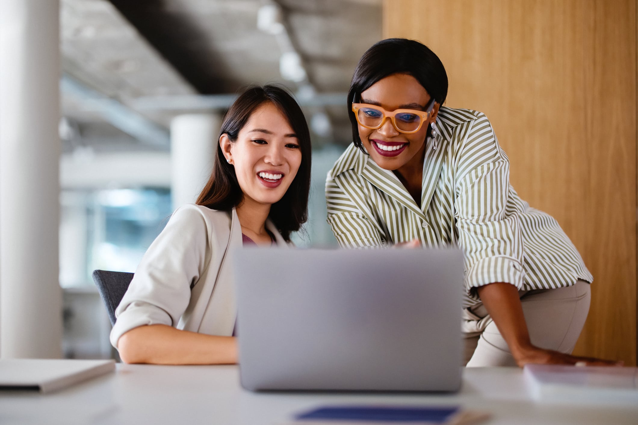 Two professional women sitting together at a desk, collaborating on a laptop in an office setting