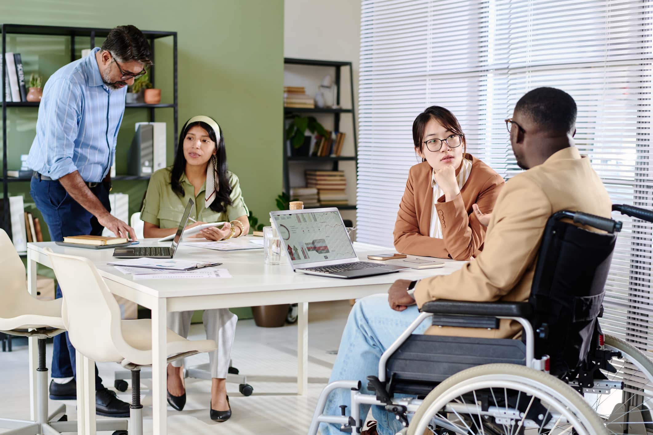 Group of colleagues, including a wheelchair user, collaborating around laptops during an office meeting
