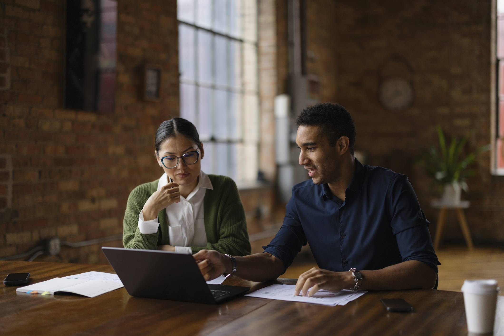 Two coworkers are sitting at a desk working on a laptop together.