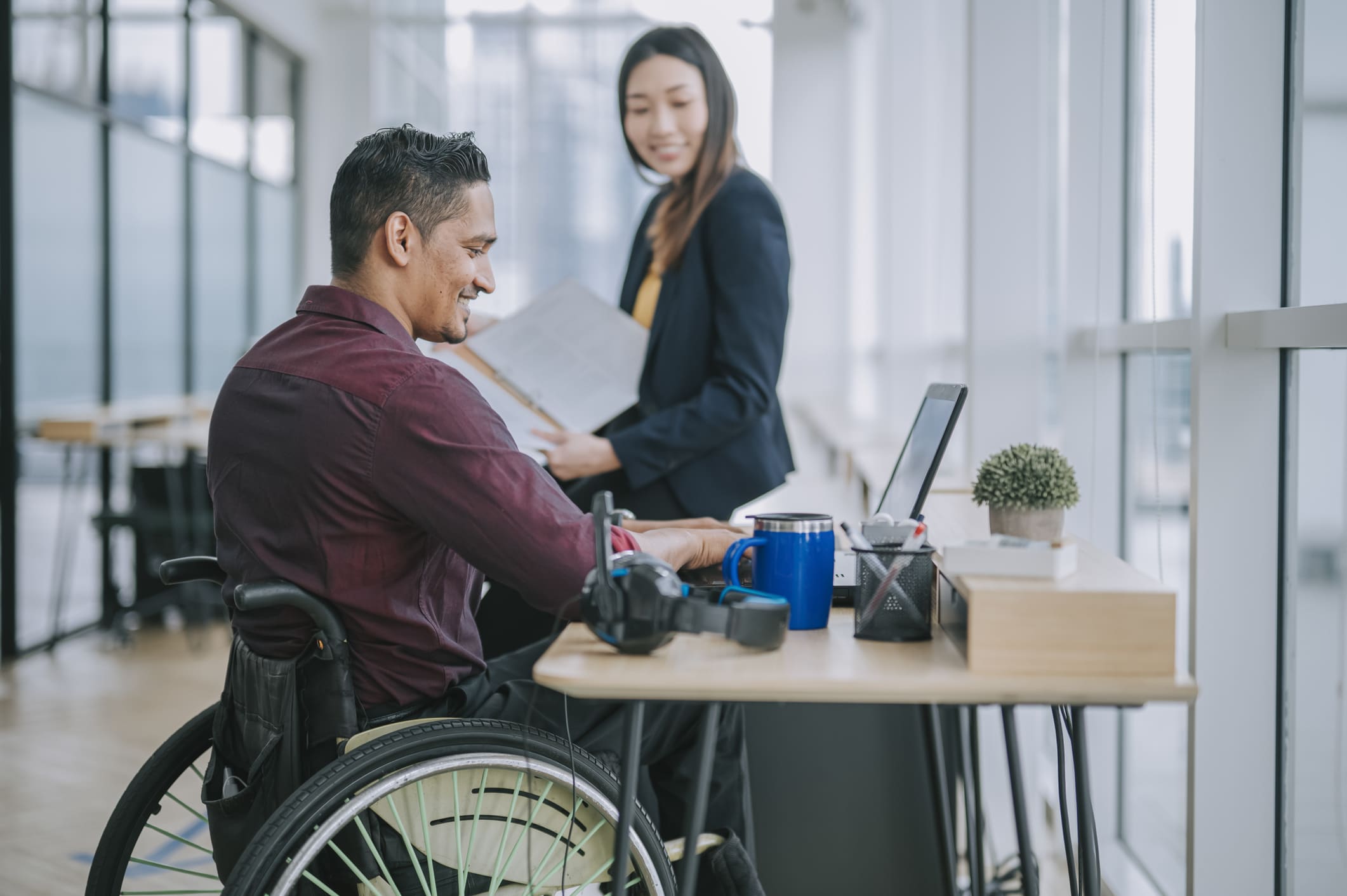 Two colleagues having a cheerful conversation in an office; one is using a wheelchair and working on a laptop.